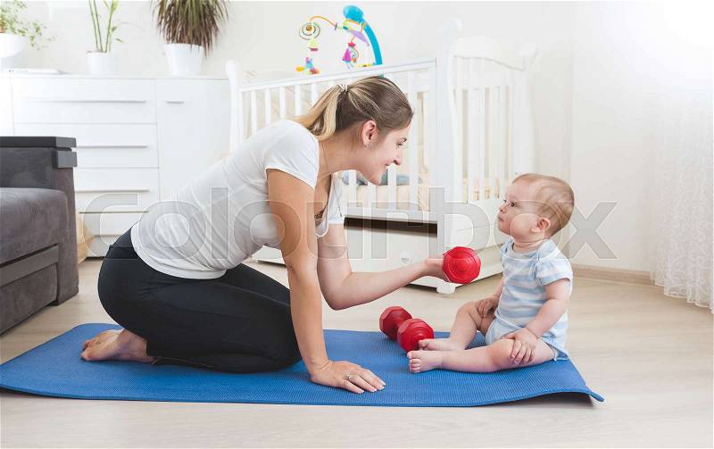 Beautiful smiling mother doing physical exercise with her baby on floor at living room, stock photo