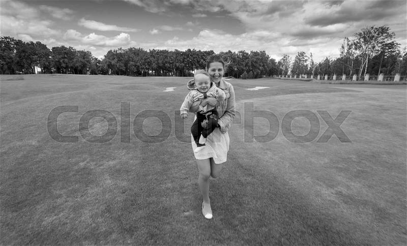 Black and white image of happy young mother running on grass at park with her 9 months old baby boy on hands, stock photo