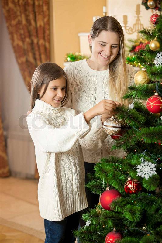 Cute girl helping mother to decorate Christmas tree at living room, stock photo