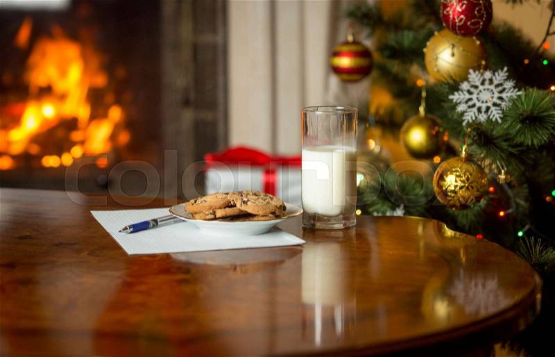 Closeup image of treats and letter to Santa on wooden table next Christmas tree and burning fireplace, stock photo