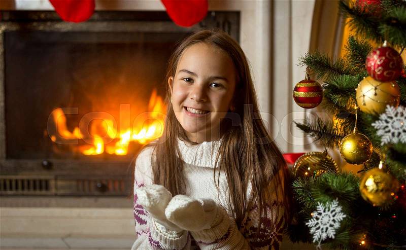 Portrait of happy smiling girl in sweater and gloves posing at burning fireplace and decorated Christmas tree, stock photo