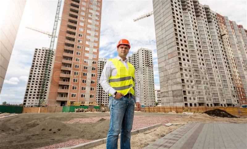 Image of young engineer in hardhat and safety vest posing against buildings under construction, stock photo