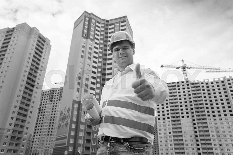Black and white portrait of smiling successful construction engineer posing on building site and showing thumb up, stock photo
