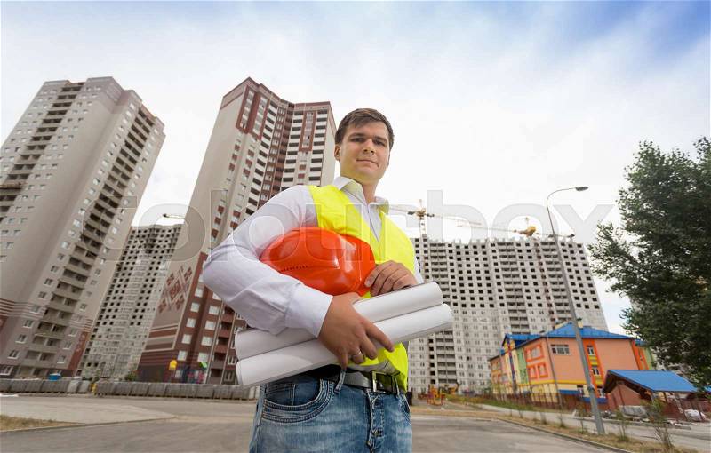 Portrait of young construction engineer in safety vest holding hardhat, stock photo