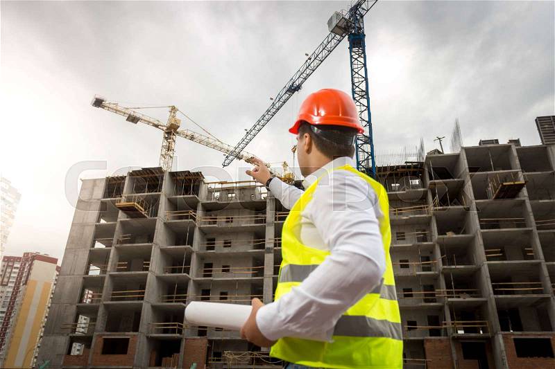 Portrait of young architect holding blueprints and pointing at building under construction, stock photo