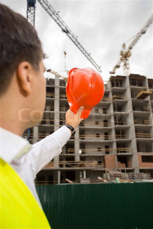 View from back on construction engineer pointing at building site with red hardhat, stock photo