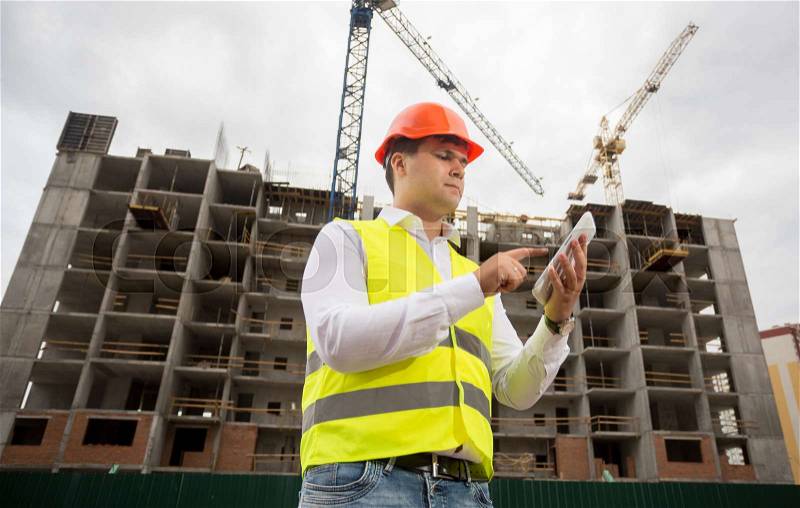 Portrait of young architect in hardhat and safety vest using digital tablet, stock photo