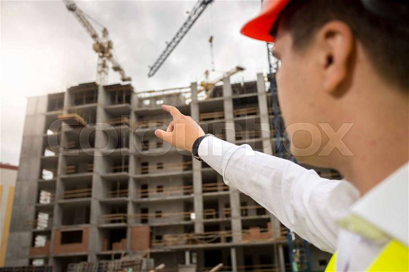 Closeup image of construction engineer pointing with hand at building under construction, stock photo