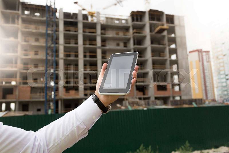 Closeup image of young male hand holding digital tablet with building site on background, stock photo
