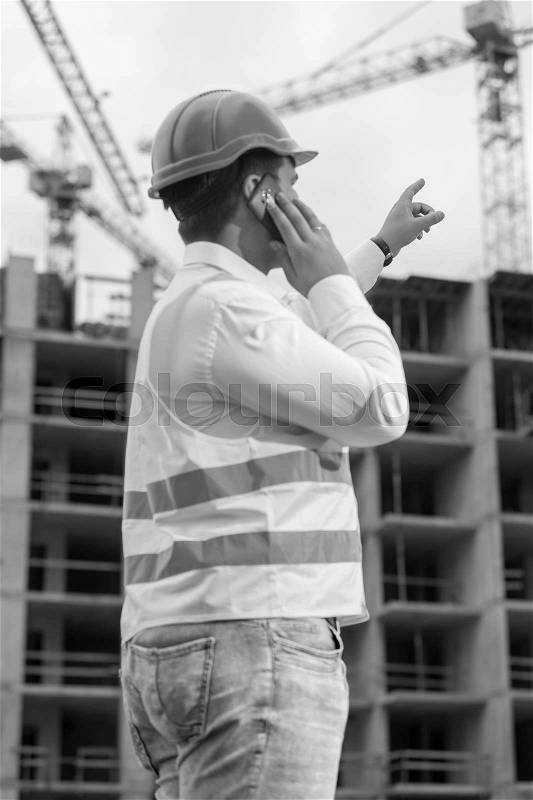 Black anf white rear view portrait of construction engineer talking by phone and pointing at building site, stock photo
