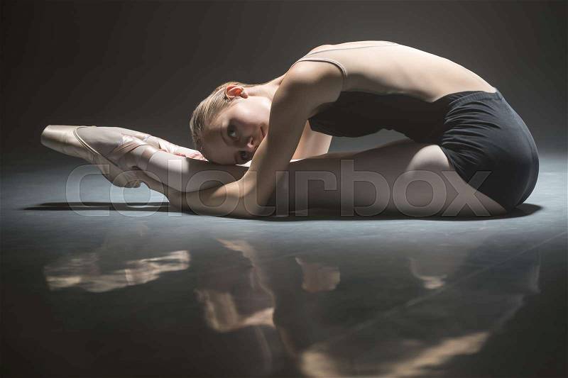 Young ballerina leaned to her feet and grabs their hands while sitting on the floor. She dressed in the black leotard. She is reflected on the floor surface. Light falls down on her back. Shoot in a low key, stock photo