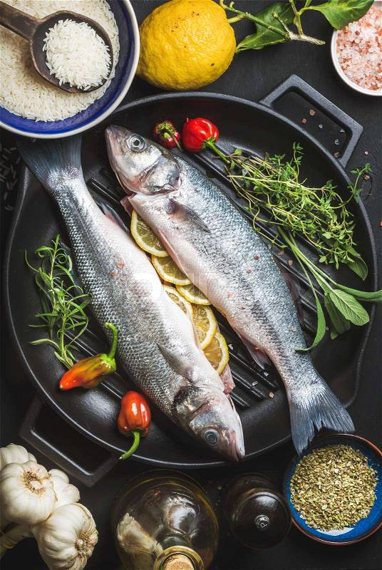Ingredients for cookig healthy fish dinner. Raw uncooked seabass fish with rice, lemon, herbs and spices on black grilling iron pan over dark background, top view,, stock photo
