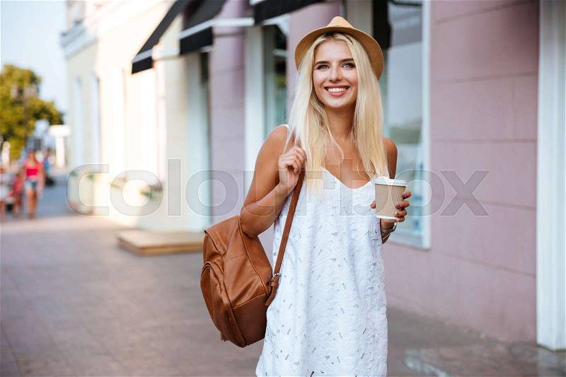 Smiling cheerful blonde girl in white dress holding take away cup outdoors, stock photo