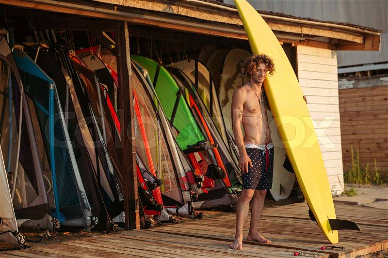 Full length of handsome curly man surfer standing and holding yellow surfing board, stock photo