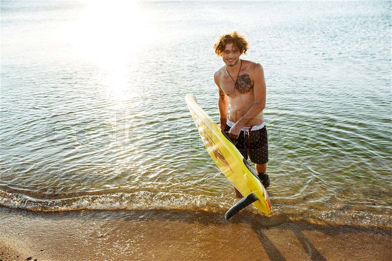 Smiling young curly man holding his surf board while standing in the ocean at the beach, stock photo