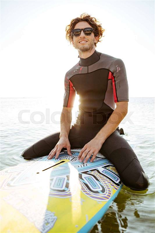 Smiling young surfer man in eyeglasses sitting on surf board in ocean and looking away, stock photo