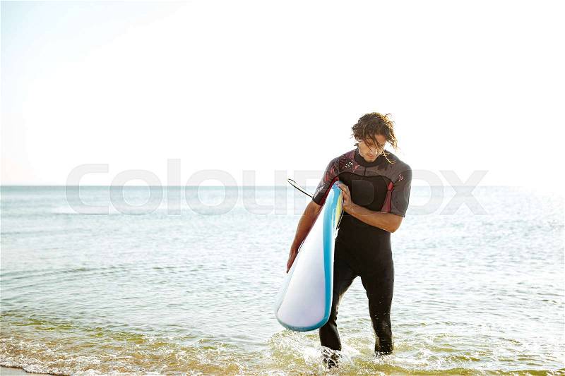 Surfer man in swimsuit walking out of the water with surf board in his hands, stock photo