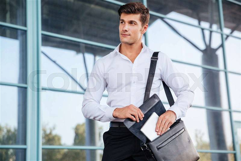 Handsome young businessman taking pc tablet from his bag while standing near business center, stock photo