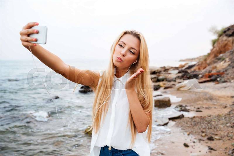 Young smiling beautiful woman sending air kiss and making selfie while standing on the rocky beach, stock photo