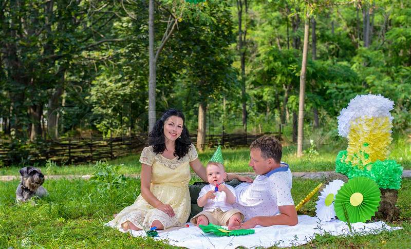 Young parents celebrating their kids first birthday relaxing outdoors in a park on rug with their little son below colorful decorations, stock photo