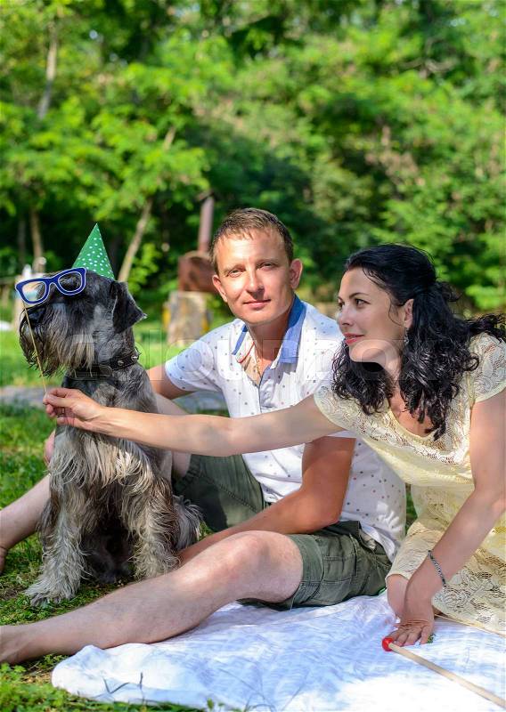 Young couple with a dog in fancy dress wearing a green party hat and photo booth glasses relaxing on a lawn in a lush park on a summer day, stock photo