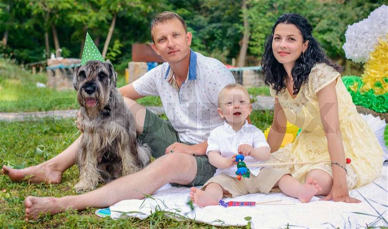Young couple with a dog in fancy dress wearing a green party hat and photo booth glasses relaxing on a lawn in a lush park on a summer day, stock photo