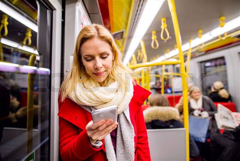 Beautiful young blond woman in red coat in subway train, holding a smart phone, texting, stock photo