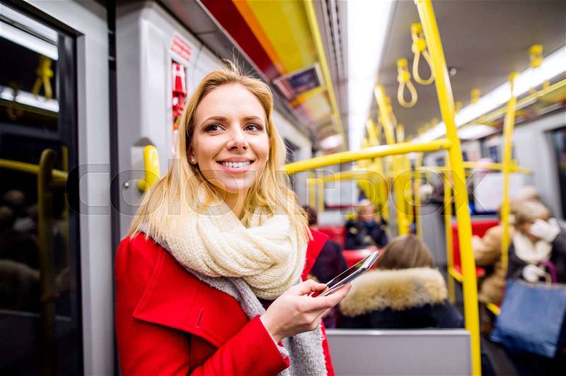 Beautiful young blond woman in red coat in subway train, holding a smart phone, texting, stock photo