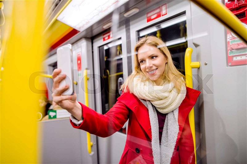 Beautiful young blond woman in red coat in subway train, holding a smart phone, taking selfie, stock photo