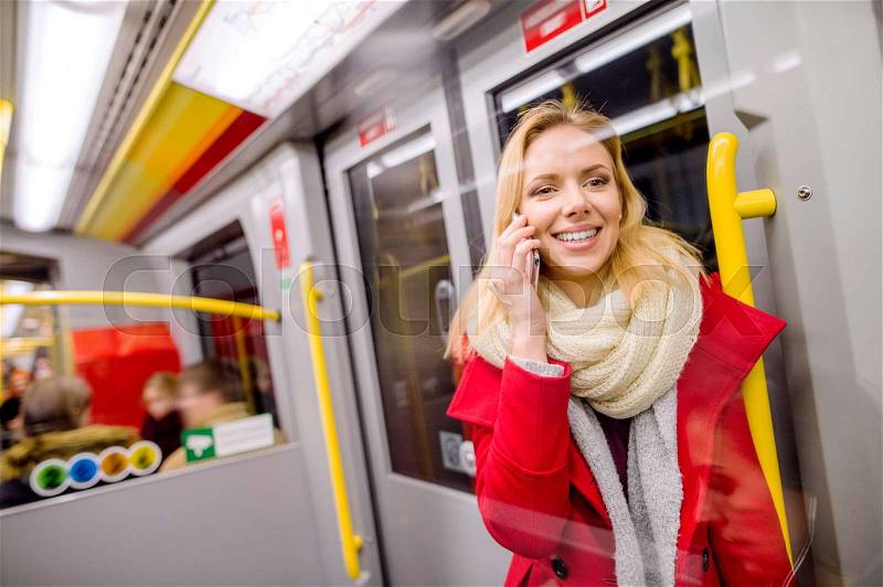 Beautiful young blond woman in red coat in subway train, holding a smart phone, making a phone call, stock photo