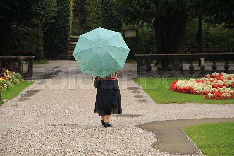The solitary lady walks under her umbrella in the rain along many rain puddles in the blooming park in the rainy summer, stock photo