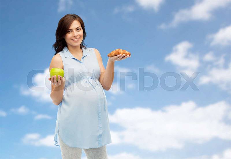 Pregnancy, healthy eating, junk food and people concept - happy pregnant woman choosing between green apple and croissant over blue sky and clouds background, stock photo