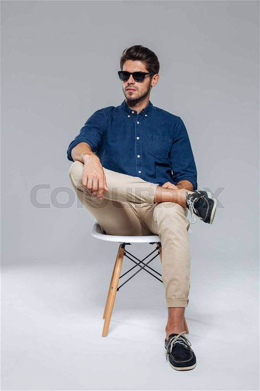 Handsome relaxed man in suglasses sitting on the chair over gray background, stock photo