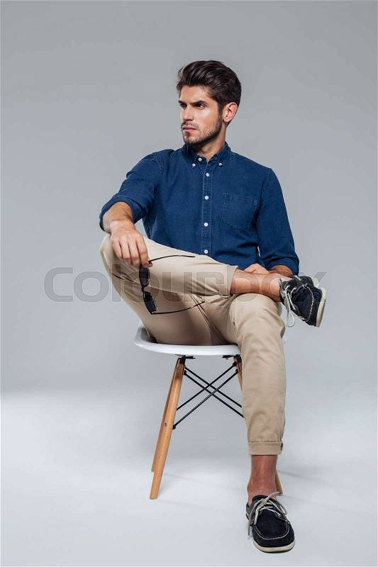 Handsome relaxed man holding suglasses and sitting on the chair over gray background, stock photo