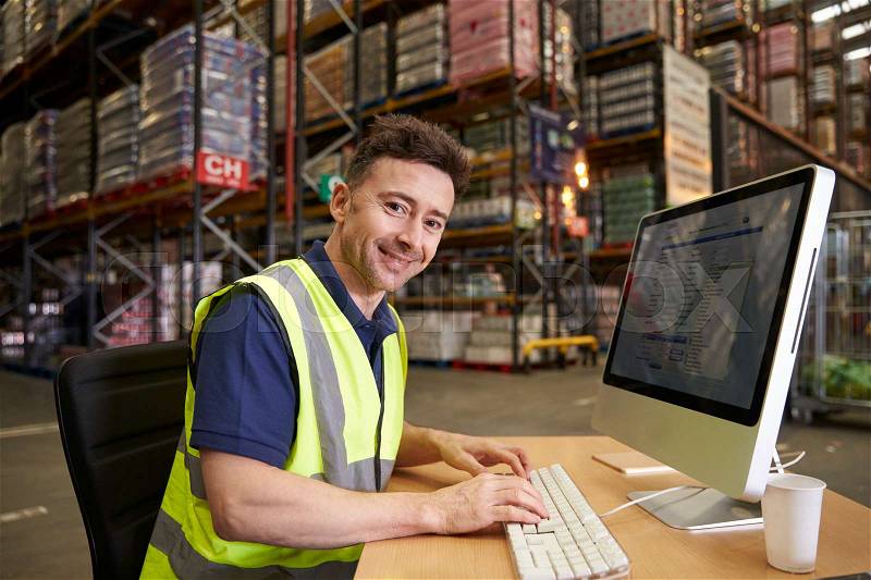 Man at computer in on-site warehouse office looks to camera, stock photo