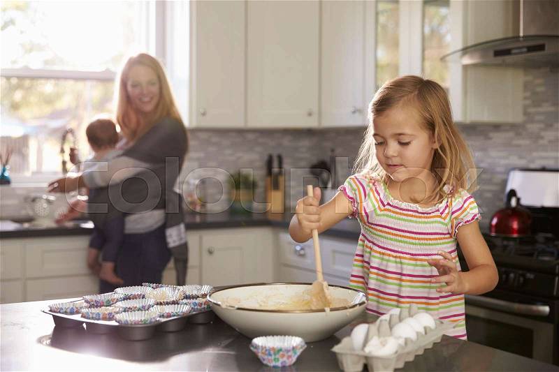 Young girl prepares cake mix, mum and baby in the background, stock photo