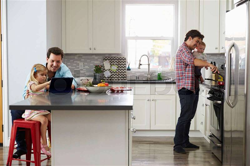 Girl uses tablet in kitchen with dad, while other dad cooks, stock photo