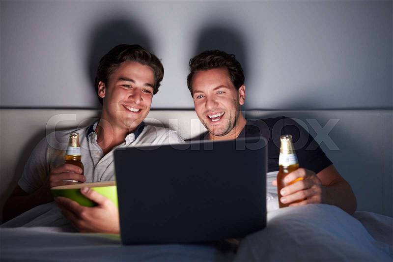 Male couple laughing in bed, drinking and watching a laptop, stock photo