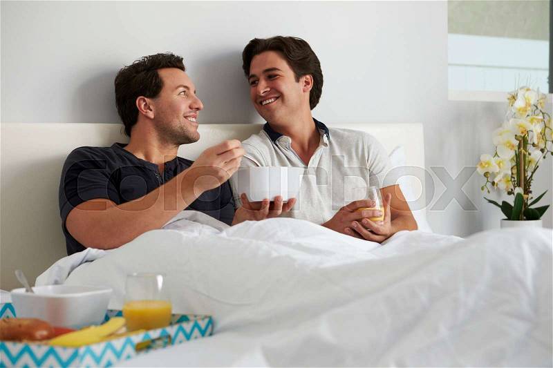 Male gay couple relax in bed eating, looking at each other, stock photo