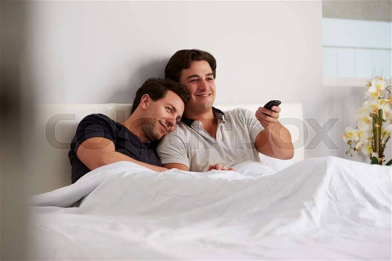 Male gay couple relax in bed together watching TV, stock photo