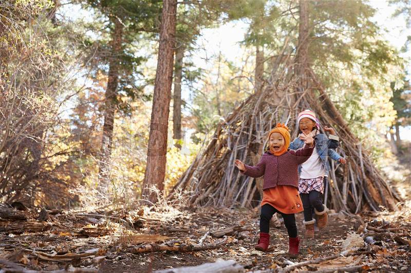 Two girls play outside shelter made of branches in a forest, stock photo