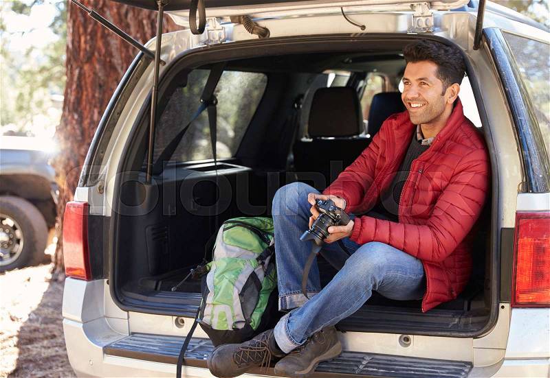Man relaxing in open back of car holding camera, stock photo