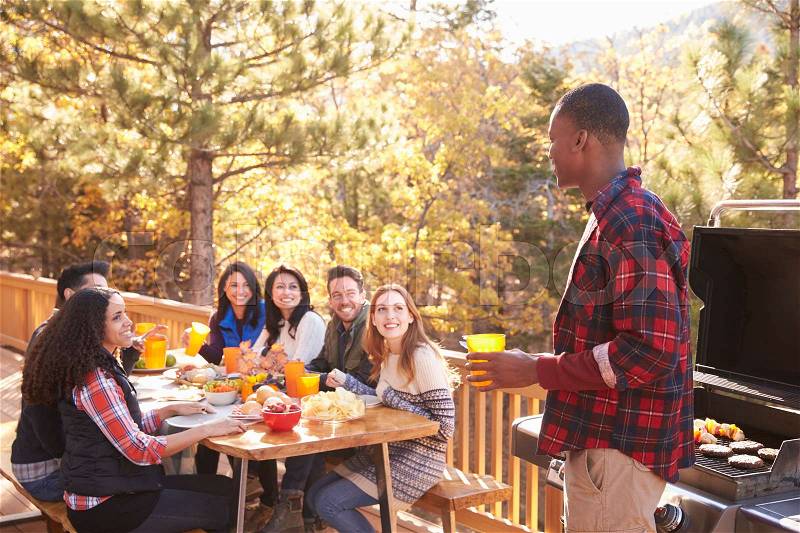 Man barbecuing on a deck talks to friends sitting at a table, stock photo