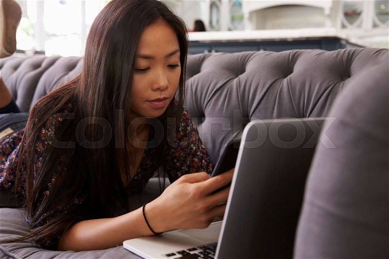 Woman Lying On Sofa At Home Using Mobile Phone And Laptop, stock photo