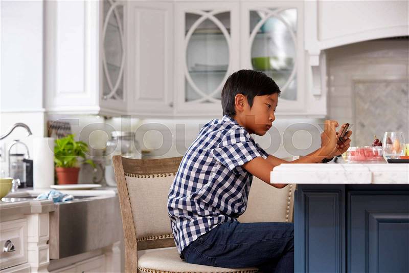 Young Asian Boy Playing Game On Mobile Device In Kitchen, stock photo