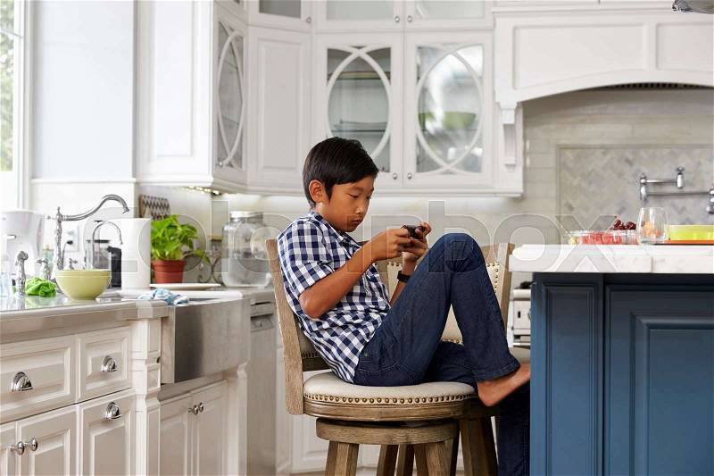 Young Asian Boy Playing Game On Mobile Device In Kitchen, stock photo