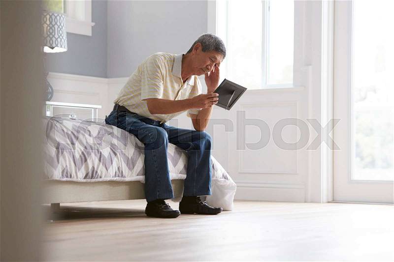 Unhappy Senior Man Sitting On Bed Looking At Photo Frame, stock photo