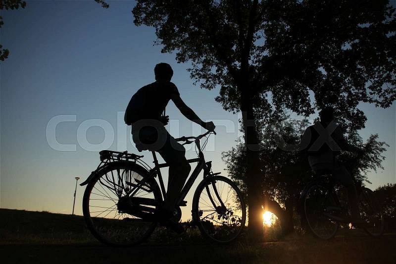 Two boys in silhouettes, one is visible and the other under the tree are biking in the wonderful park in the village at sunset in the summer, stock photo