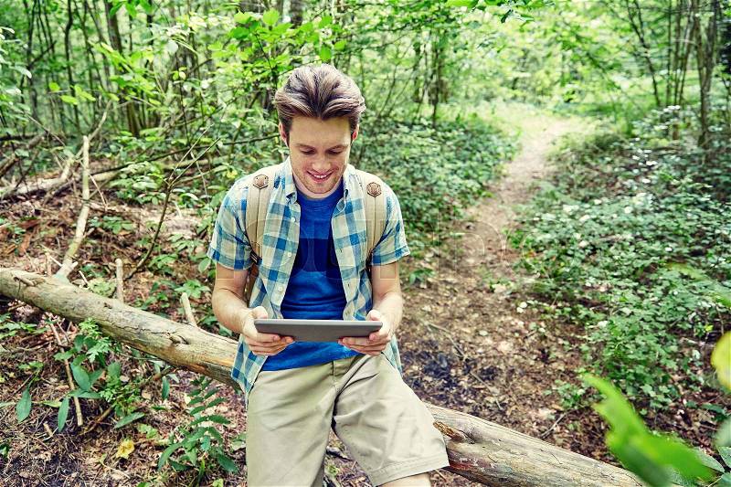 Adventure, travel, tourism, hike and people concept - happy young man with backpack and tablet pc computer sitting on fallen tree trunk in woods, stock photo