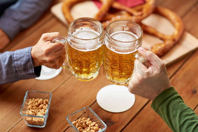 People, leisure and drinks concept - close up of male hands clinking beer mugs and pretzels at bar or pub, stock photo